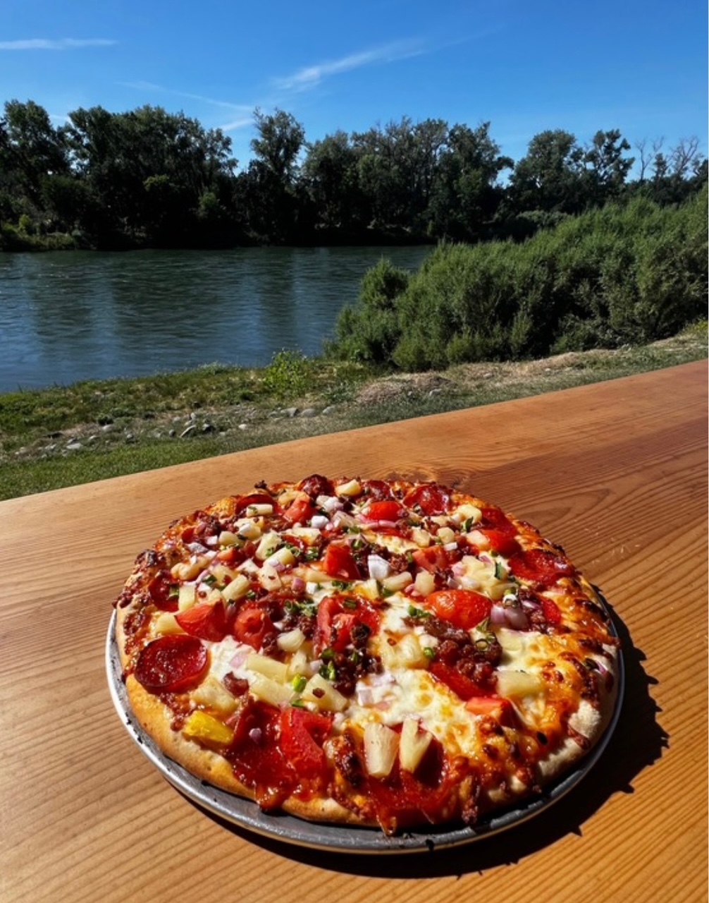 A pepperoni, pineapple and tomato pizza on a metal pan resting on a wooden rail above the Sacramento River.